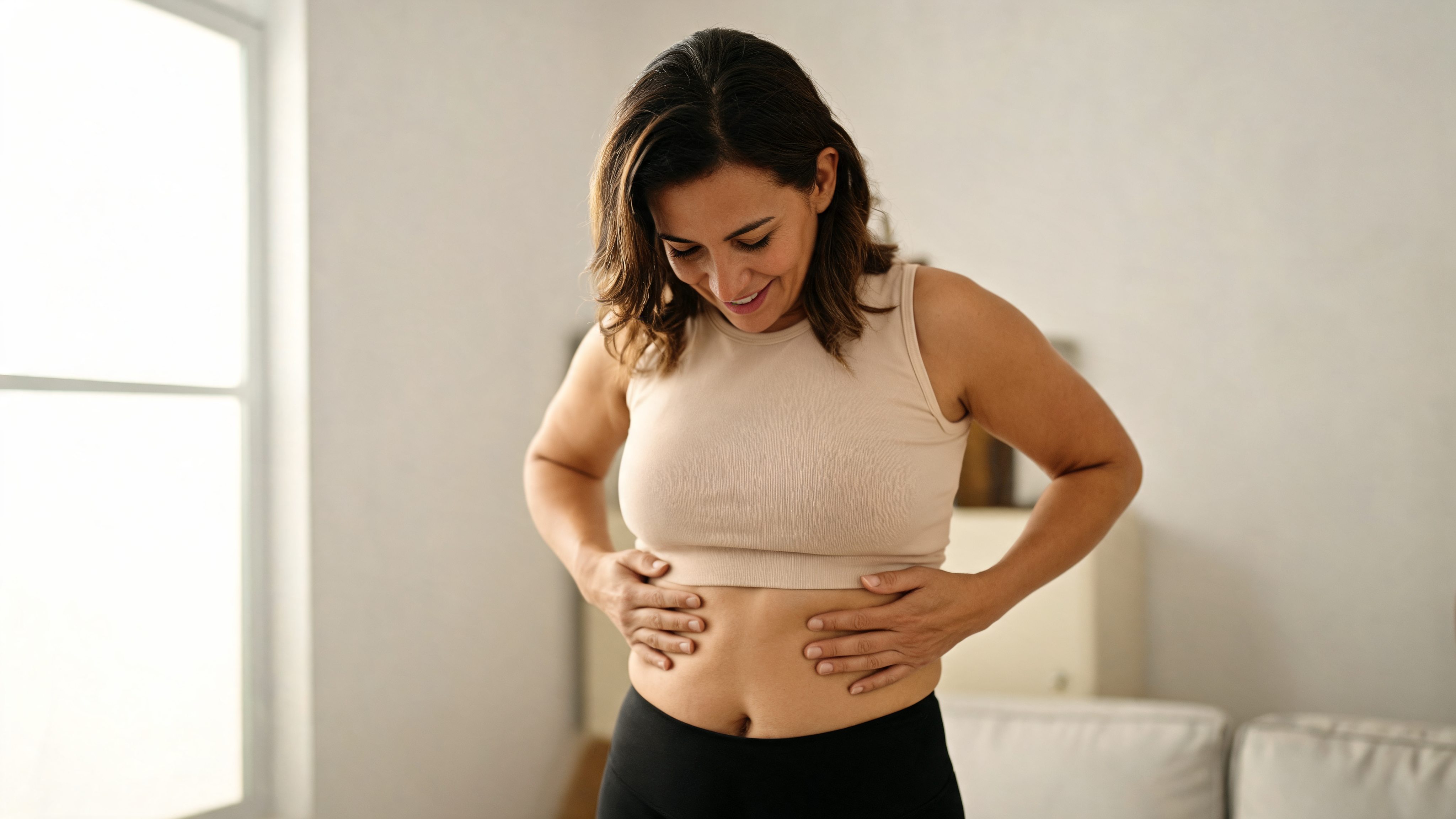 Mulher sorrindo com as mãos sobre a barriga, em roupa de treino, na sala de casa